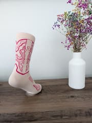 The pink and cream boot sock displayed on a wooden shelf beside a vase of dried flowers.